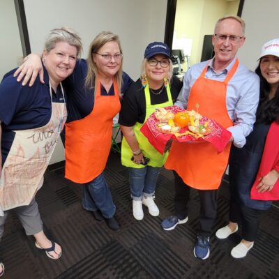 A group of five people smiling and posing, holding a tray of food. They are participating in a chili cook-off team building activity. Three of the participants wear bright aprons, one in orange and one in lime green. The man in the center is holding a red tray with an arrangement of colorful vegetables and food items.