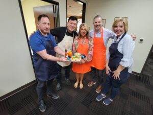 A group of five people standing together, smiling and holding a dish they prepared during a team building chili cook-off event. The group is standing indoors, with each person wearing an apron of different colors (blue, orange, and white). They are posing proudly with their food creation, likely showcasing it for the competition. The atmosphere is lighthearted, and the participants appear happy and engaged.