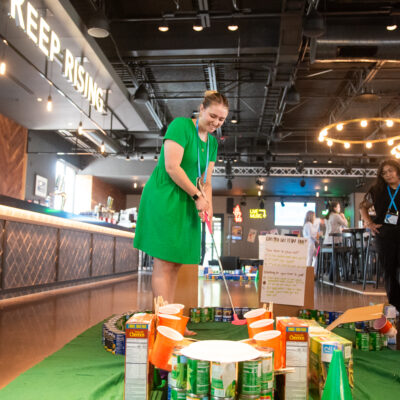 A participant plays on a creatively designed mini golf course made from canned goods and food packages during a 'Build Your Own Mini Golf Course' team building event. The event takes place in a lively venue, combining fun, teamwork, and charity, with the food items used for the course being donated to a local food pantry afterward.