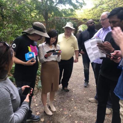 A group of team members gathered in a garden, reviewing clues and maps during an outdoor scavenger hunt. Each person is focused on their materials, including instant cameras and printed instructions, as they collaborate to solve challenges.