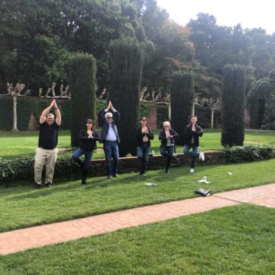 A group of team members standing in a landscaped garden, striking synchronized playful poses with hands above their heads during an outdoor team building event. They stand in front of neatly trimmed hedges and greenery, creating a lighthearted and engaging moment in a scenic, natural environment.