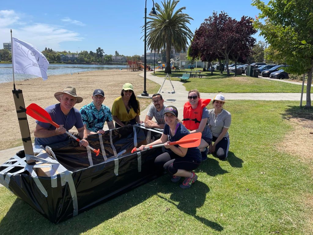 Team posing proudly with their completed cardboard boat and paddles during the Build-A-Boat team building activity by the waterfront.