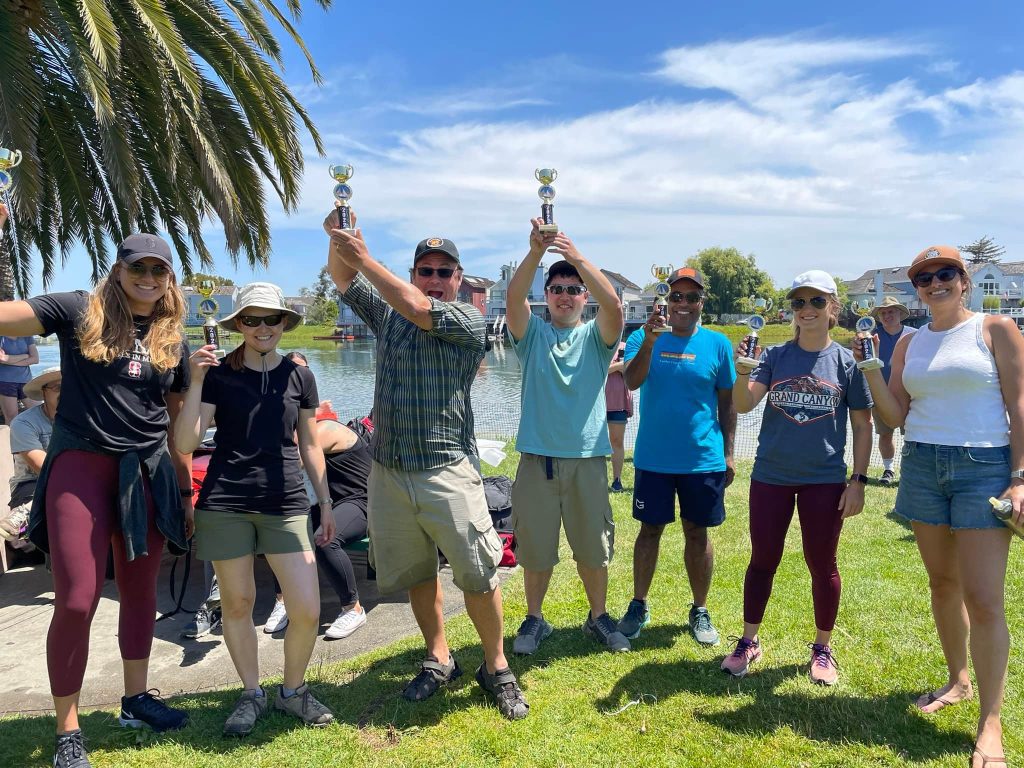 Team members celebrating with trophies after successfully completing the Build-A-Boat team building challenge by the water.