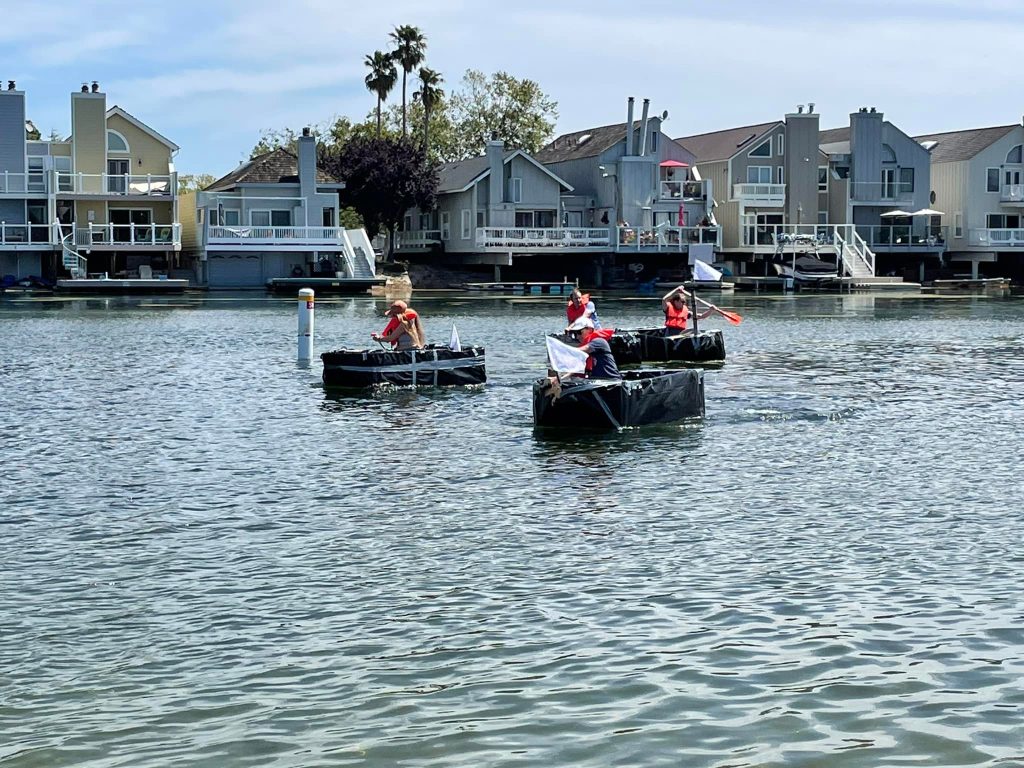 Teams paddling their cardboard boats across the lake during the Build-A-Boat team building challenge.