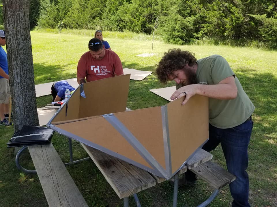 Team member assembling the cardboard structure of a boat during the Build-A-Boat team building activity in a park setting.