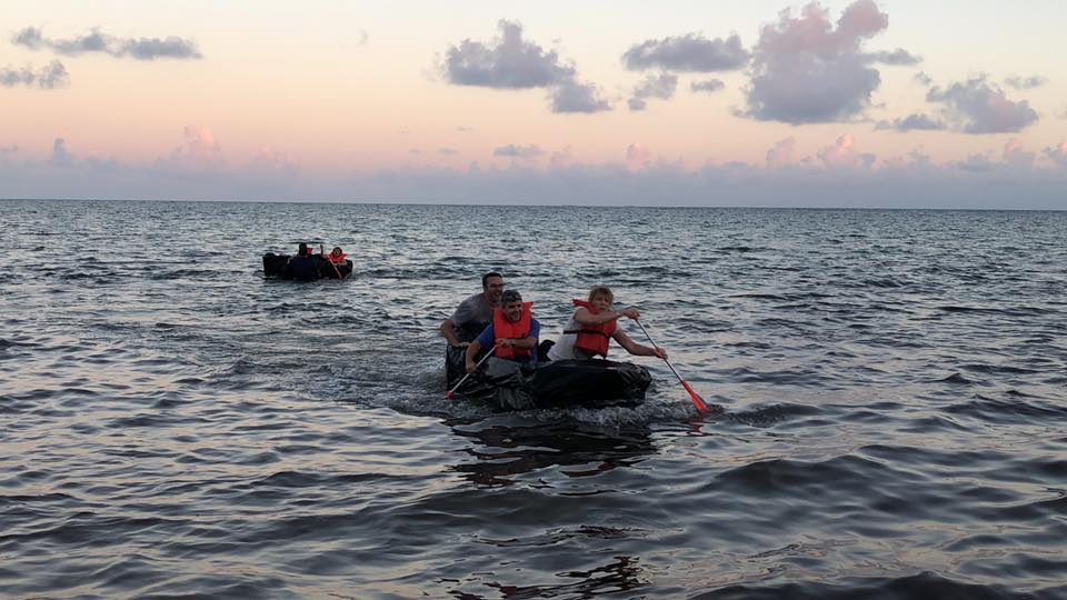 Teams paddling cardboard boats on the open water during the Build-A-Boat team building challenge at sunset.