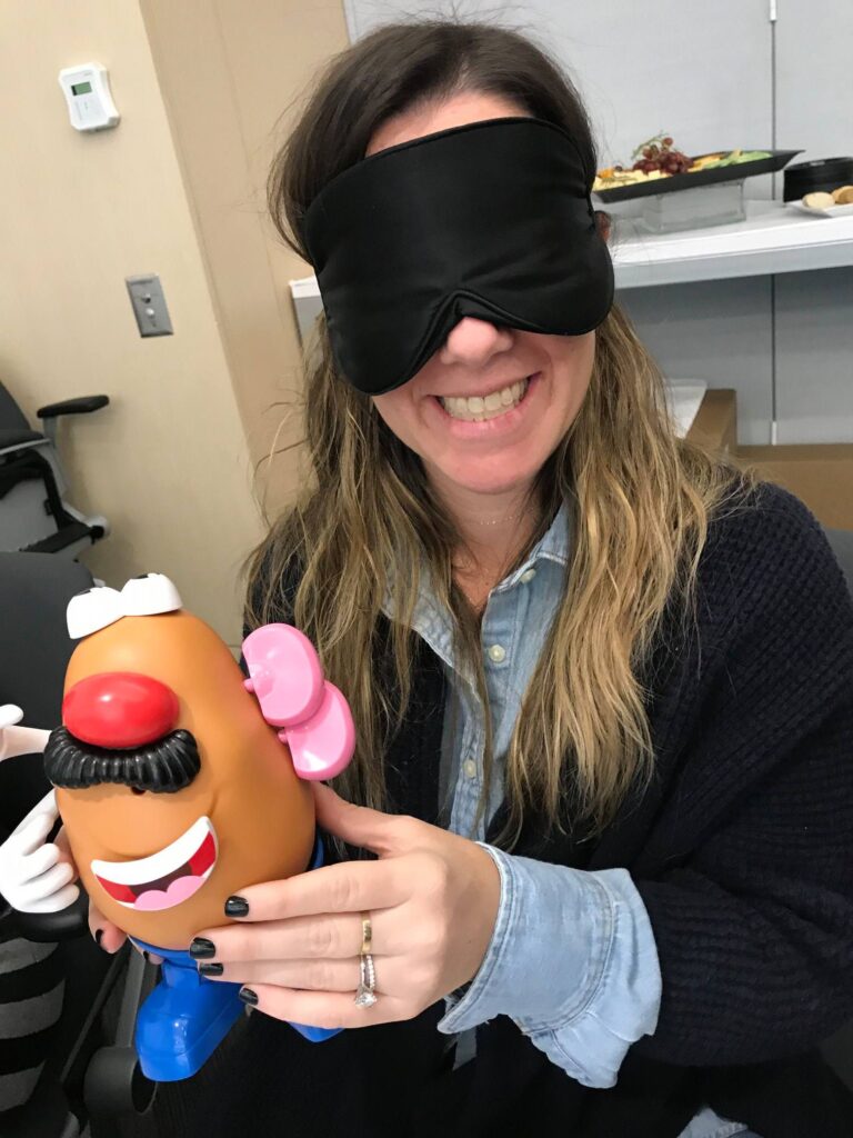 A woman smiling and holding a Mr. Potato Head toy while wearing a black blindfold. She is participating in a fun activity as part of the Toys for Tykes team building event, where participants assemble a toy blindfolded as a challenge.