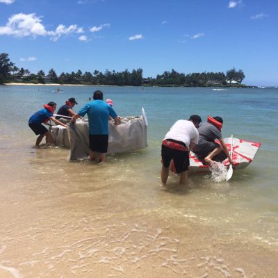 Teams launching their handmade cardboard boats into the water during the Build-A-Boat team building challenge on a sunny beach.