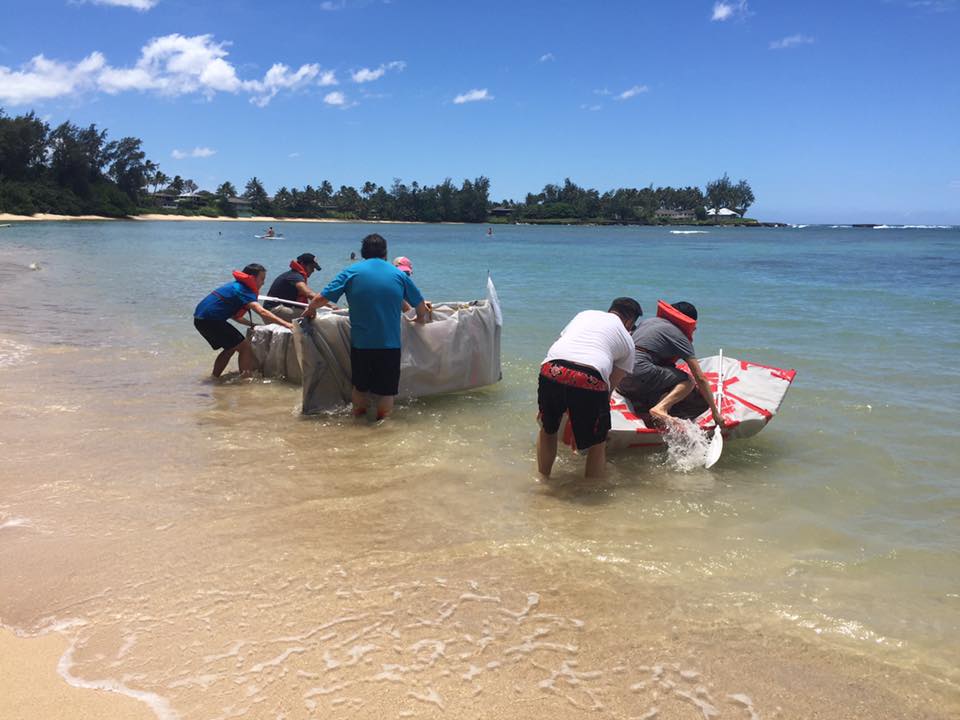 Teams launching their handmade cardboard boats into the water during the Build-A-Boat team building challenge on a sunny beach.