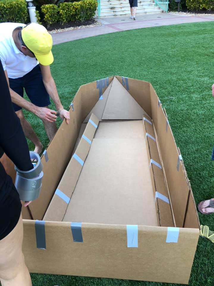 Team members constructing a cardboard boat framework during the Build-A-Boat team building challenge on a grassy outdoor space.