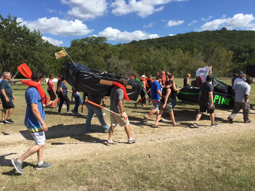 Teams carry their creatively designed cardboard boats during the Build-A-Boat team building event on a sunny day.