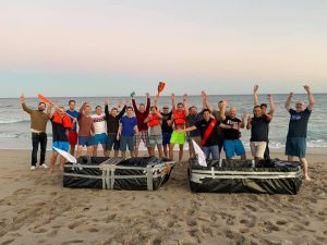 "Team members celebrate on the beach with their completed cardboard boats during the Build-A-Boat team building challenge.