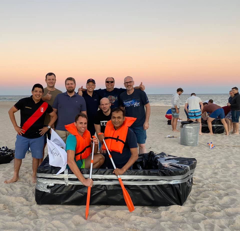 Team poses with their completed cardboard boat on the beach during the Build-A-Boat team building event at sunset.