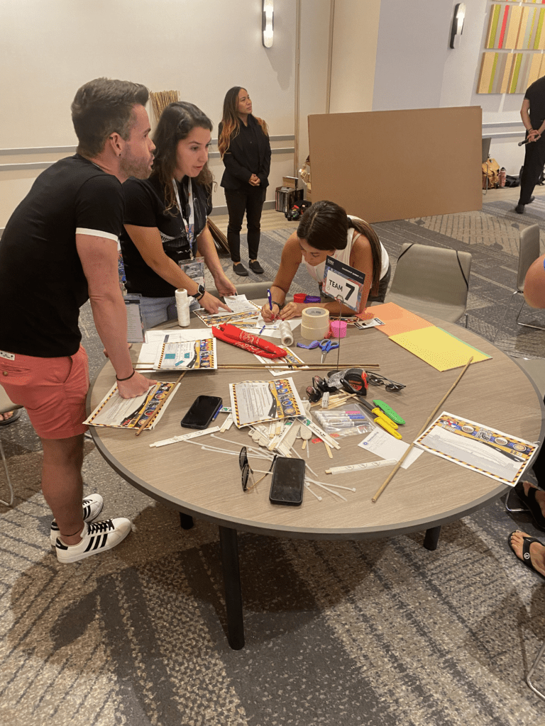 Team members collaborate at a table filled with supplies, working on their bridge design for the Bridging the Divide team building activity.