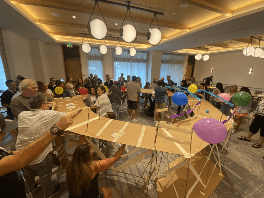 Teams collaborate to construct a large bridge decorated with balloons during the Bridging the Divide team building activity.