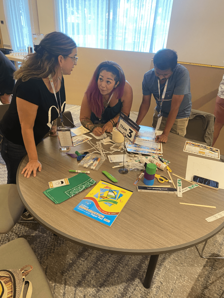 Team 3 members brainstorm and plan their bridge design at a table covered with materials during the Bridging the Divide team building activity.