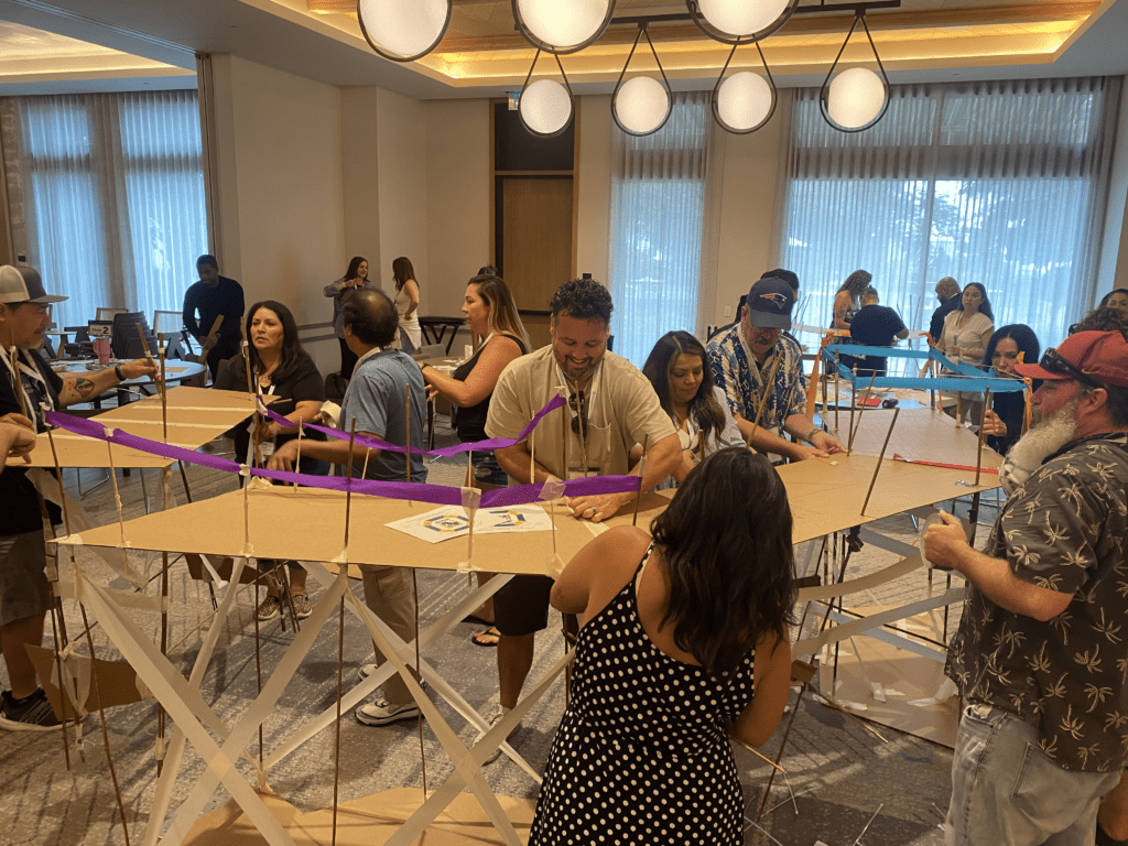 Teams work together to build a large bridge structure using cardboard and colorful materials during the Bridging the Divide team building activity.