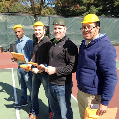 Four team members wearing yellow safety helmets, standing together outdoors on a tennis court, smiling and holding team building materials during the 'Catapult to Success' team building activity.