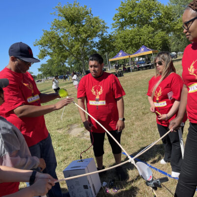 A team of participants in red shirts working together outdoors on constructing a catapult during the Catapult to Success team building activity, showcasing collaboration, creativity, and strategic planning.