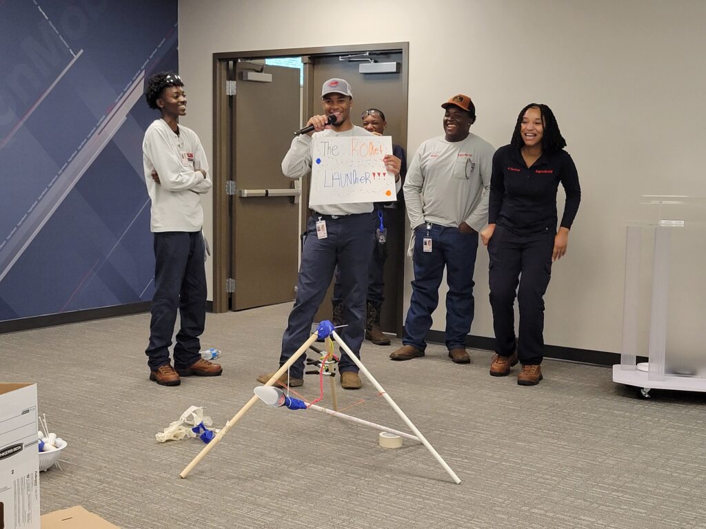 A team presenting their catapult design called 'The Rocket Launcher' during an indoor segment of the Catapult to Success team building activity, showcasing creativity, teamwork, and presentation skills.