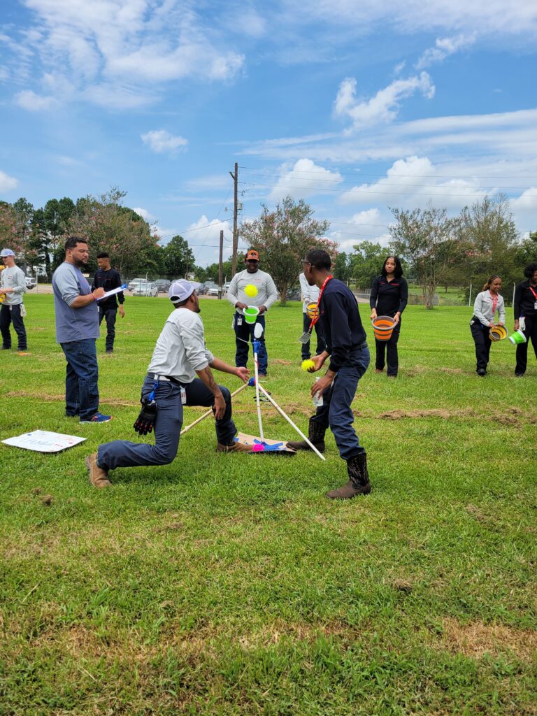 Participants outdoors engaging in a team building exercise with a catapult setup, preparing for a launch while other team members observe and strategize, demonstrating teamwork, collaboration, and competitive spirit in the Catapult to Success activity.