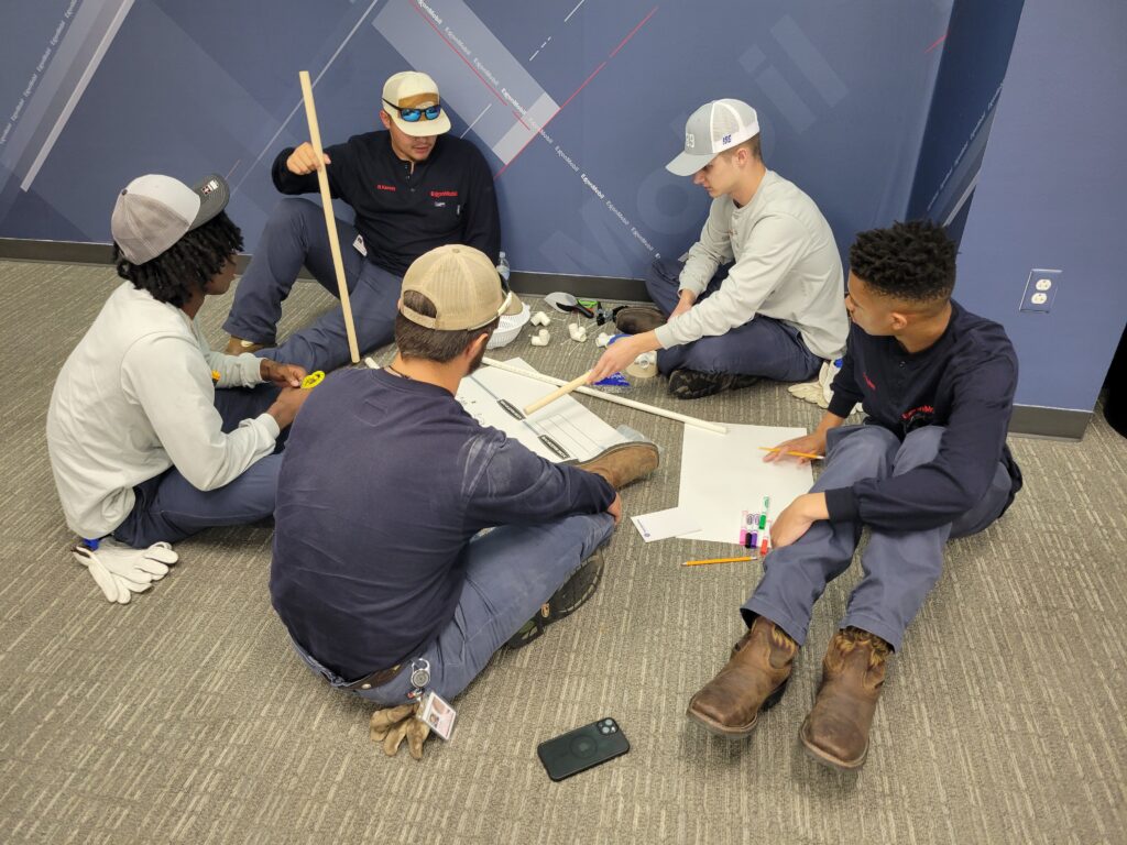 A group of participants sitting on the floor indoors, discussing and planning their catapult design during the Catapult to Success team building activity, emphasizing teamwork, collaboration, and strategic thinking.