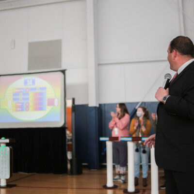 A game show host holding a microphone and looking at a large screen displaying the game board, while participants cheer and clap during the Survey Says team building activity in a gymnasium setting.