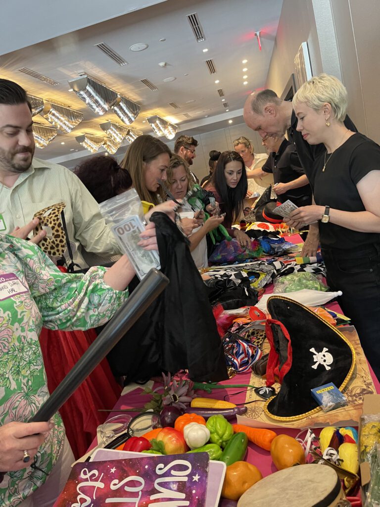 A group of people enthusiastically selecting costumes and props from a table filled with various items like pirate hats, colorful fabrics, and accessories during a team building activity focused on creativity and fun role-playing.