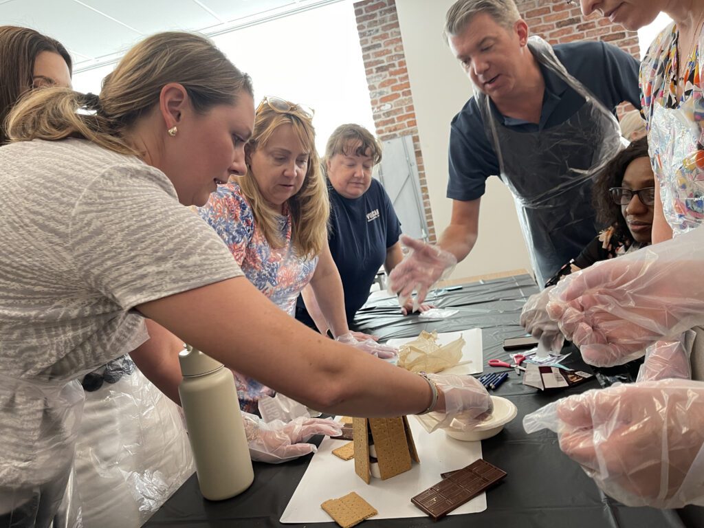 Participants in The Chocolate Challenge team building event work together to construct a structure out of graham crackers and chocolate, demonstrating collaboration and creative problem-solving.