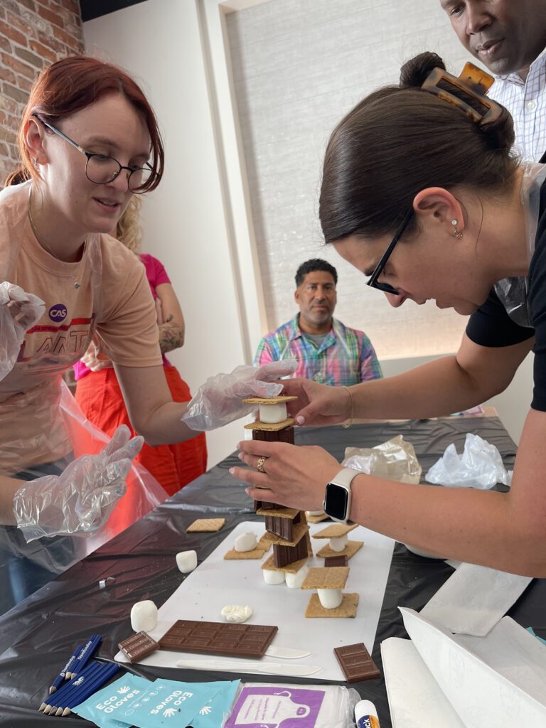 Team members collaborate to build a tall structure out of chocolate, graham crackers, and marshmallows during The Chocolate Challenge team building event, showcasing teamwork and creativity.