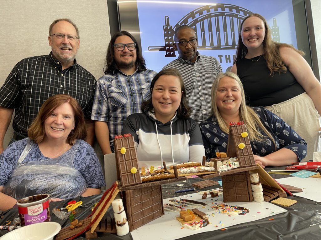 A team of seven poses proudly behind their creative chocolate bridge during The Chocolate Challenge team building event. The structure, made entirely of chocolate, includes detailed elements like wafers and sprinkles. Smiling participants showcase their teamwork and edible engineering skills.