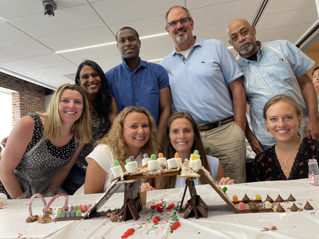 A team proudly poses behind their edible chocolate bridge creation during The Chocolate Challenge team building event. The bridge is decorated with candy, marshmallows, and other sweet treats, highlighting teamwork and creativity.