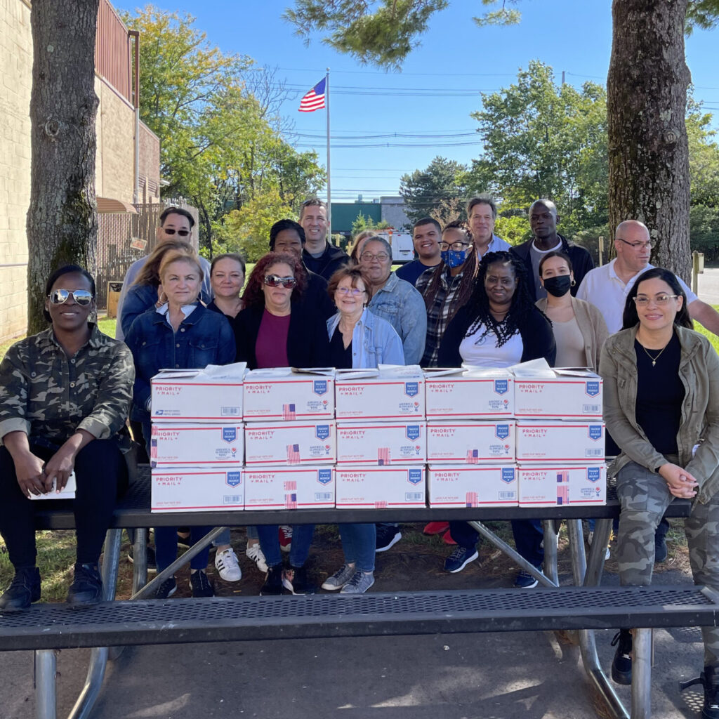 Team members gather with care packages they've assembled during an Operation Military Care team building event, ready to support U.S. military personnel stationed overseas.