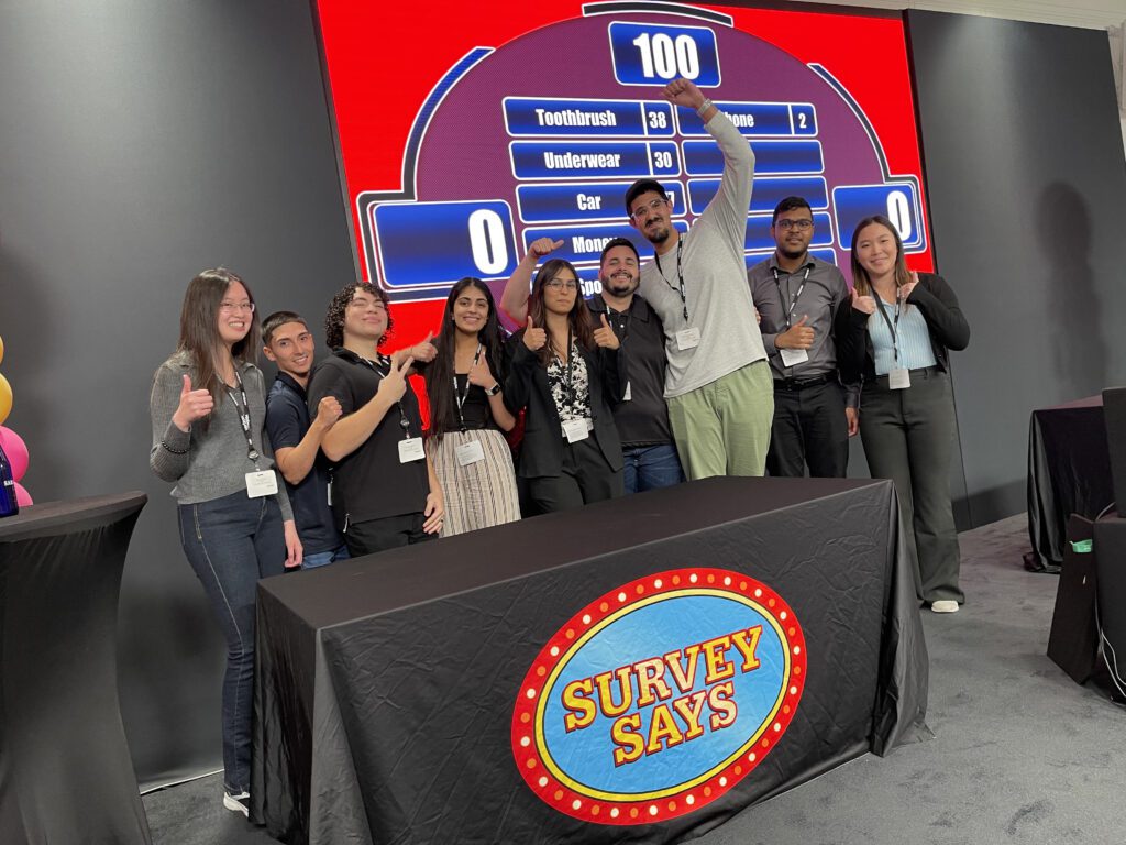 A group of participants smiling and giving thumbs up in front of a large game board during the Survey Says team-building activity, celebrating their participation in a fun, competitive, and interactive game.