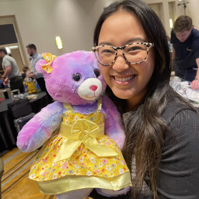 A smiling woman holds a brightly colored purple and pink teddy bear dressed in a yellow dress, as part of a charitable team building event called Team Teddy Rescue Bear.