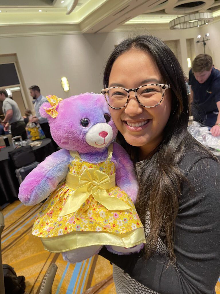 A smiling woman holds a brightly colored purple and pink teddy bear dressed in a yellow dress, as part of a charitable team building event called Team Teddy Rescue Bear.