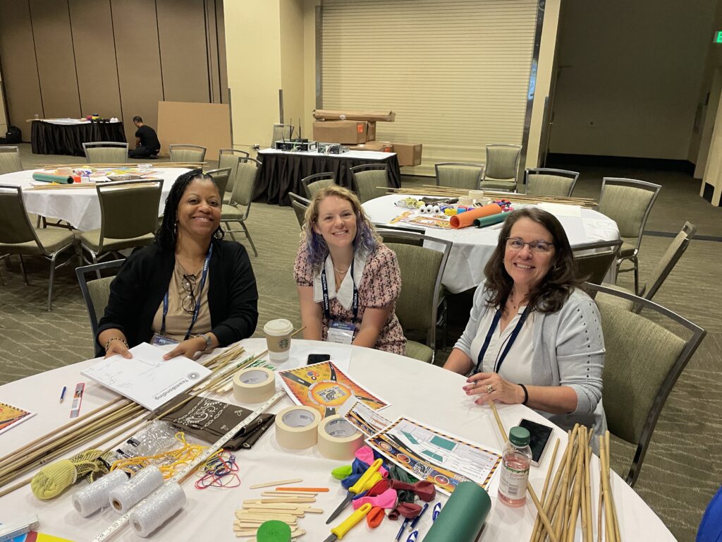 Three participants smile during the Bridging the Divide team building event, seated at a table filled with creative supplies for the bridge-building activity.