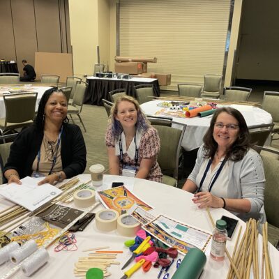 Three participants smile during the Bridging the Divide team building event, seated at a table filled with creative supplies for the bridge-building activity.