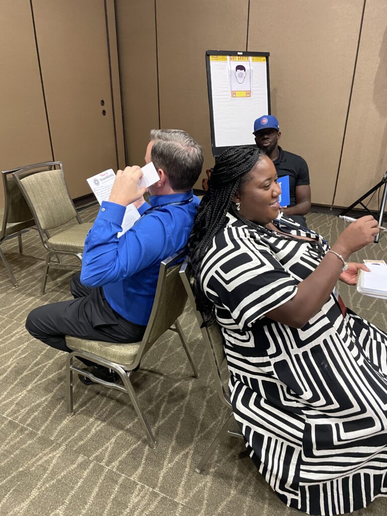 Two participants engage in a collaborative communication exercise, sitting back-to-back as part of the Bridging the Divide team building activity.