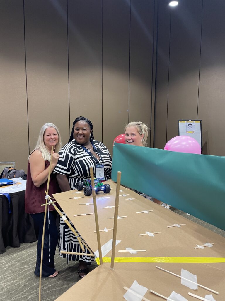 Participants smile next to their completed bridge, ready to test it with a radio-controlled car during the Bridging the Divide team building activity.