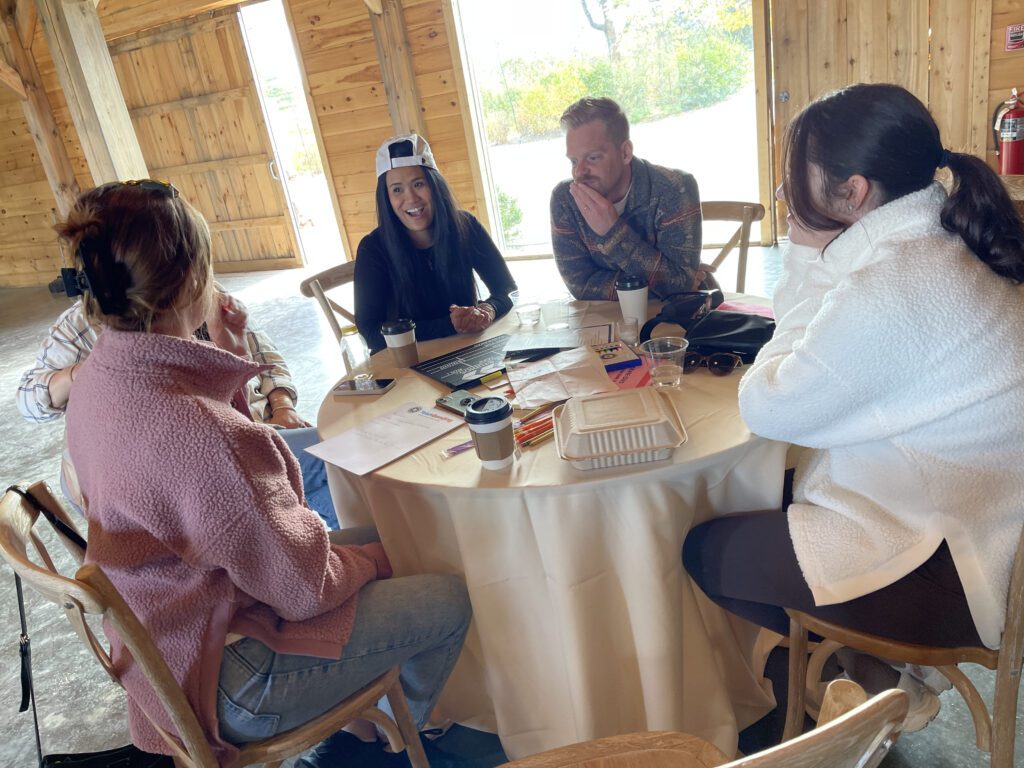 Group of four people sitting around a round table engaged in a discussion, with papers, coffee cups, and a laptop, capturing a collaborative team building moment.