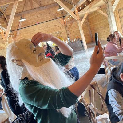 Person wearing a long white-bearded mask taking a selfie with a smartphone, preparing for the Corporate Film Festival challenge.