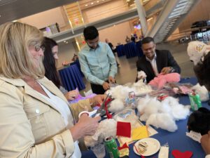 A group of participants enthusiastically engages in the Team Teddy Rescue Bear team building activity. A woman in a beige jacket is cutting fabric, while others nearby are working with teddy bear stuffing and various craft supplies on a blue table.