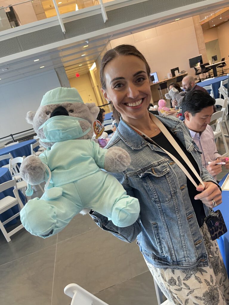 A smiling participant holds a teddy bear dressed as a doctor in light blue scrubs and a matching face mask. The bear is part of the Team Teddy Rescue Bear team building event, where participants create and dress up teddy bears for charity.