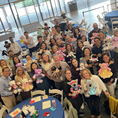 A large group of participants proudly holding their completed teddy bears at the end of the Team Teddy Rescue Bear event. The participants, arranged in a group around circular tables, smile and showcase the colorful, dressed-up teddy bears they made as part of the team building CSR event.