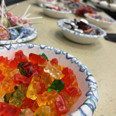 Close-up of a bowl filled with colorful gummy bears, with additional bowls of candies and lollipops visible in the background, prepared for a sweet treat station during The Chocolate Challenge team building event.