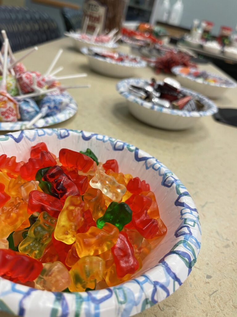 Close-up of a bowl filled with colorful gummy bears, with additional bowls of candies and lollipops visible in the background, prepared for a sweet treat station during The Chocolate Challenge team building event.