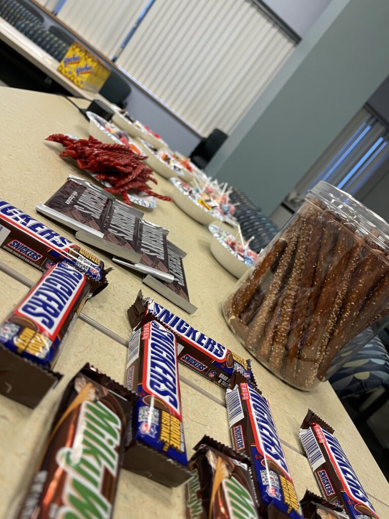 A table filled with various snacks, including candy bars, pretzels, and sweets, prepared for The Chocolate Challenge team building event, offering participants tasty materials to use in their creative constructions.