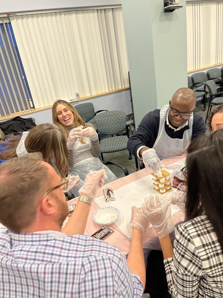 A group of colleagues works together to stack chocolate pieces during The Chocolate Challenge team building event. They are smiling and wearing aprons and gloves, showcasing their teamwork and enjoying the fun activity.