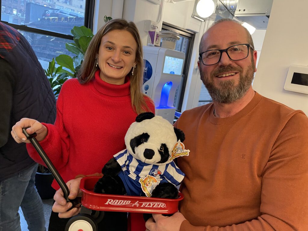 A man and a woman stand together smiling, holding a small red Radio Flyer wagon with a stuffed panda bear dressed in a blue and white striped outfit. They are participating in a charitable team building event, showing their completed project with pride and joy.